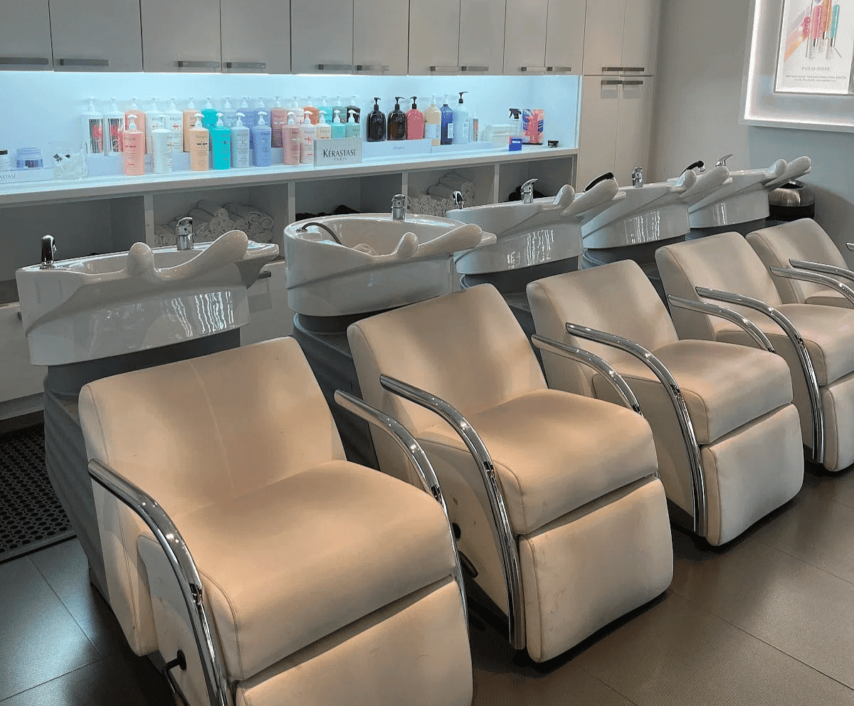 Modern salon with white chairs and sinks lined up in front of a well-stocked product shelf.